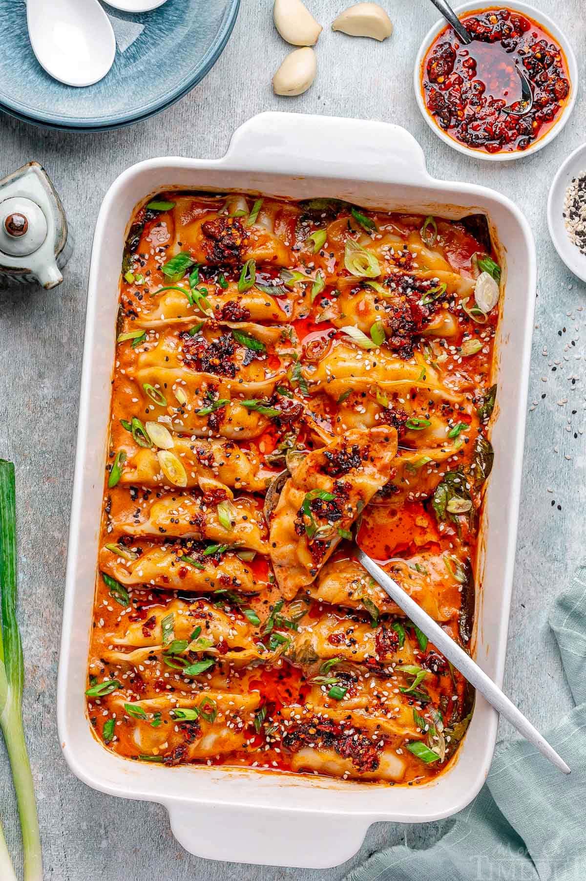 Overhead shot of viral baked dumplings in white baking dish topped with chili crips, green onions and black and white sesame seeds. Spoon in dish ready to serve the dumplings.
