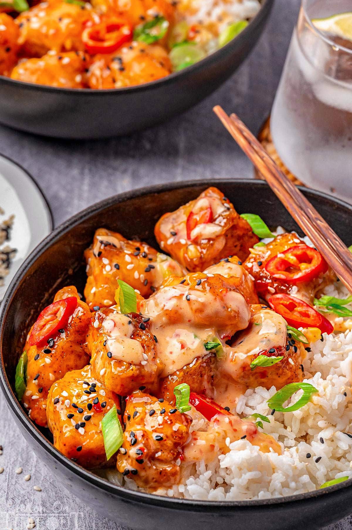 Close up shot of bang bang salmon bites in black bowl served with rice and lemon wedges. Another bowl can be seen in the background.