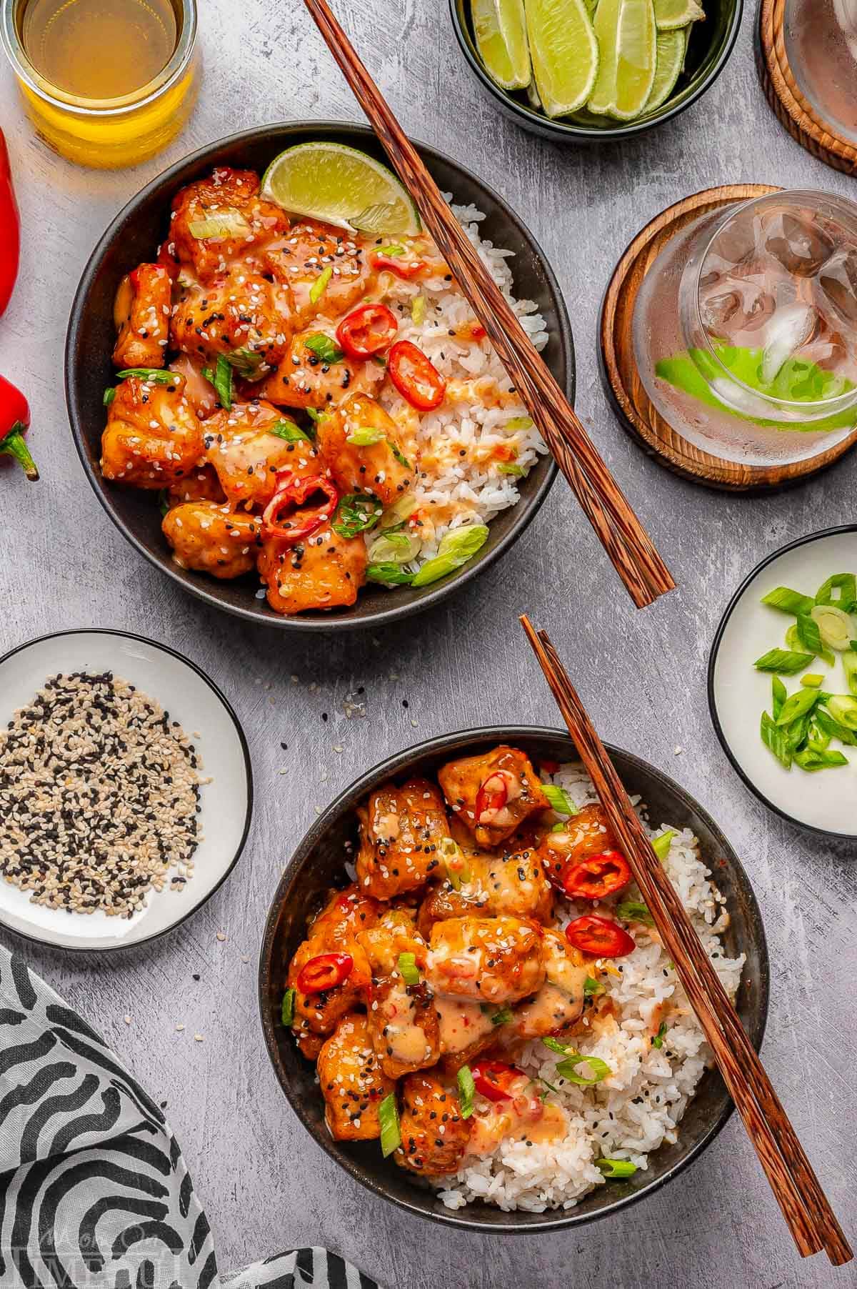 Overhead shot of two bowl of crispy bang bang salmon bites served with rice and garnishes. Chopsticks resting on edge of bowl.