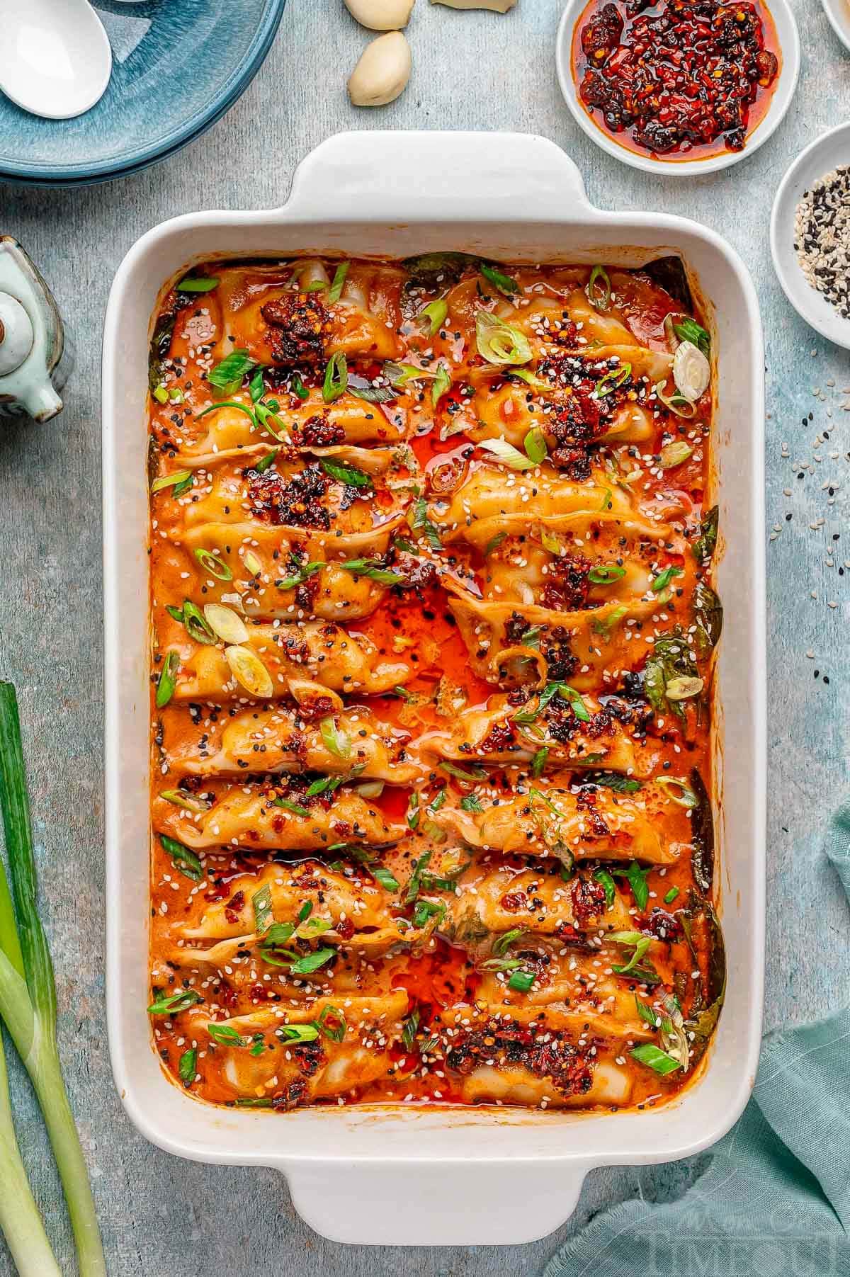 Overhead shot of viral baked dumplings in white baking dish topped with chili crips, green onions and black and white sesame seeds. Spoon in dish ready to serve the dumplings.