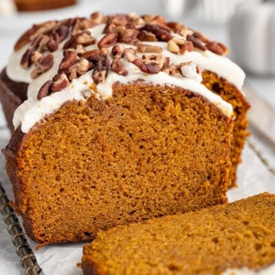 Easy pumpkin bread loaf sitting on cutting board with a few pieces sliced off. Loaf is topped with cream cheese frosting and pecans.
