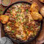 Overhead shot of french onion pot roast in cast iron skillet with cheese on top and baguette slices scattered about. Fresh parsley garnishes the dish.