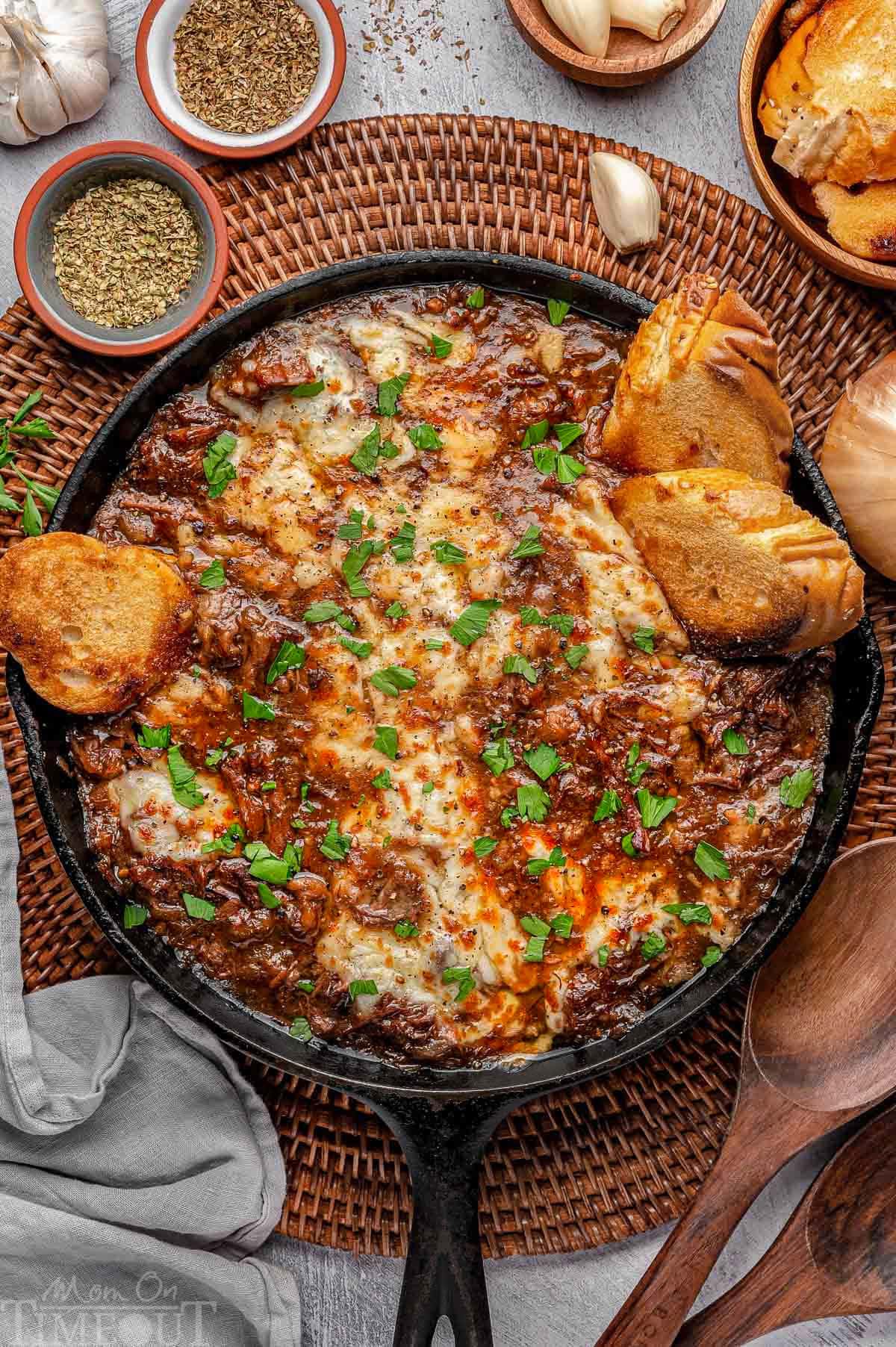 Overhead shot of french onion pot roast in cast iron skillet with cheese on top and baguette slices scattered about. Fresh parsley garnishes the dish.