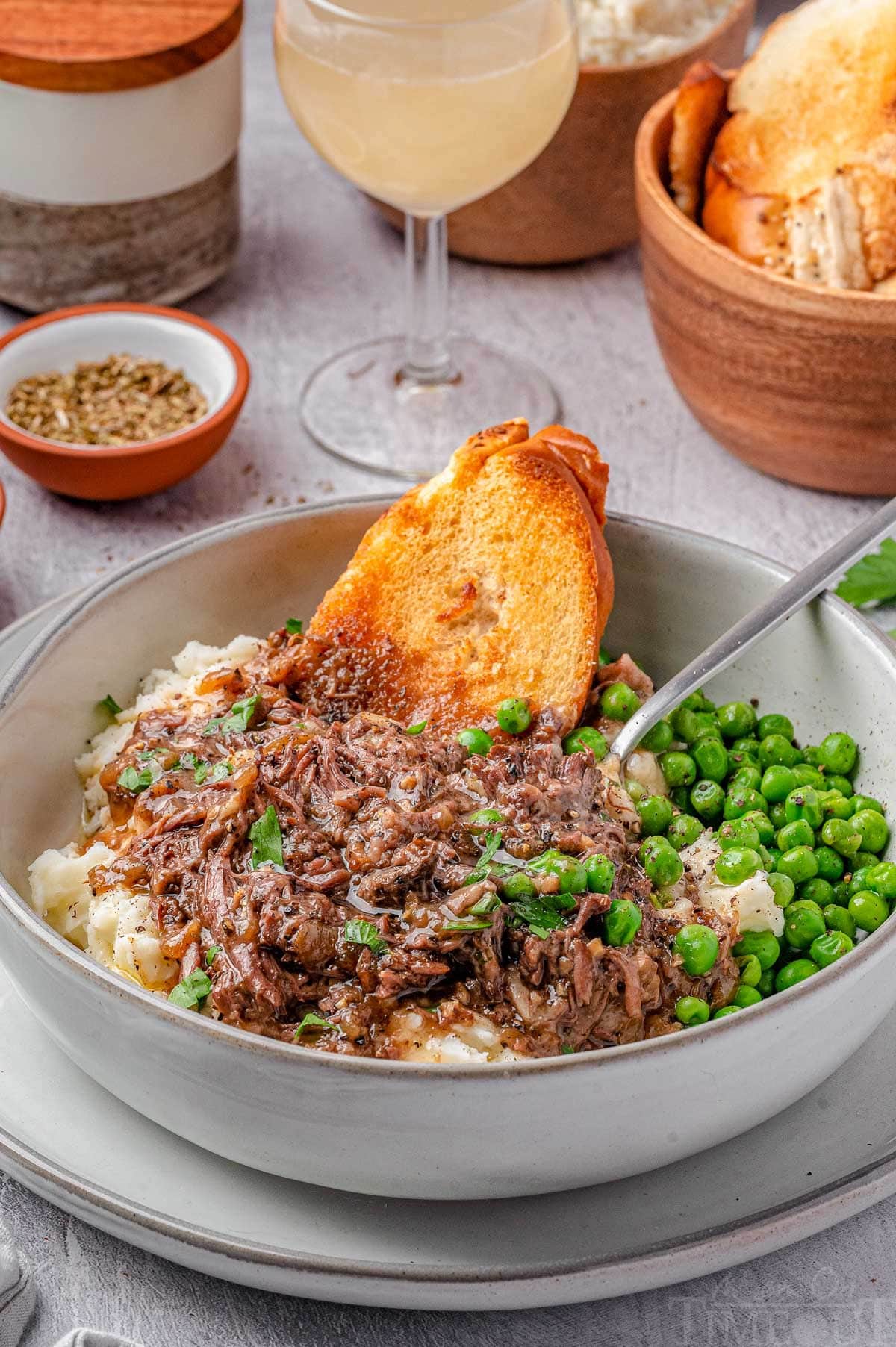 Bowl of french onion soup pot roast with mashed potatoes and peas and slices of baguette.