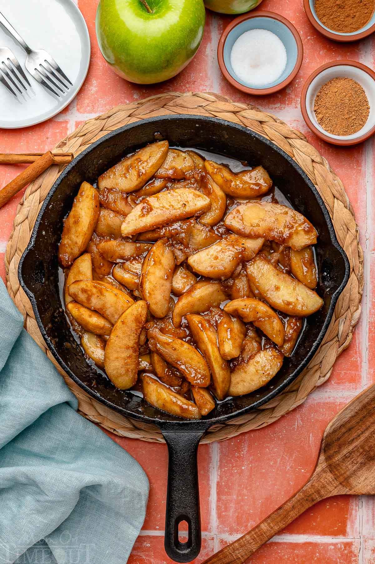 Overhead shot of fried apples in cast iron skillet. Spices and sugar in small bowls around it.
