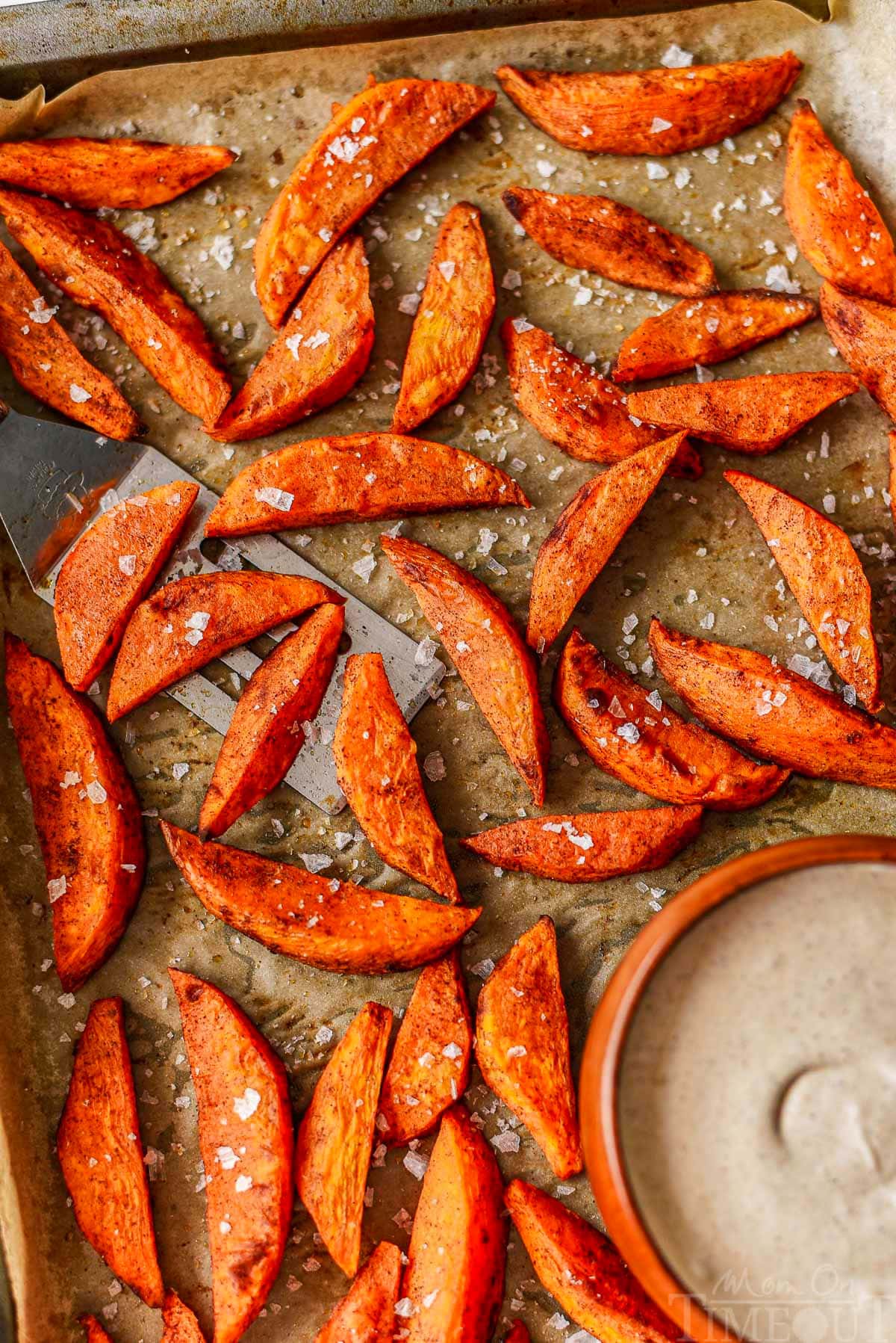 Top down look at roasted sweet potato wedges on parchment lined baking sheet with brown sugar cinnamon dip in bowl.