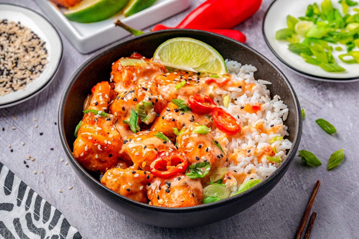 Wide shot of salmon bang bang bites in black bowl garnished with sesame seeds, green onions and lime wedges.