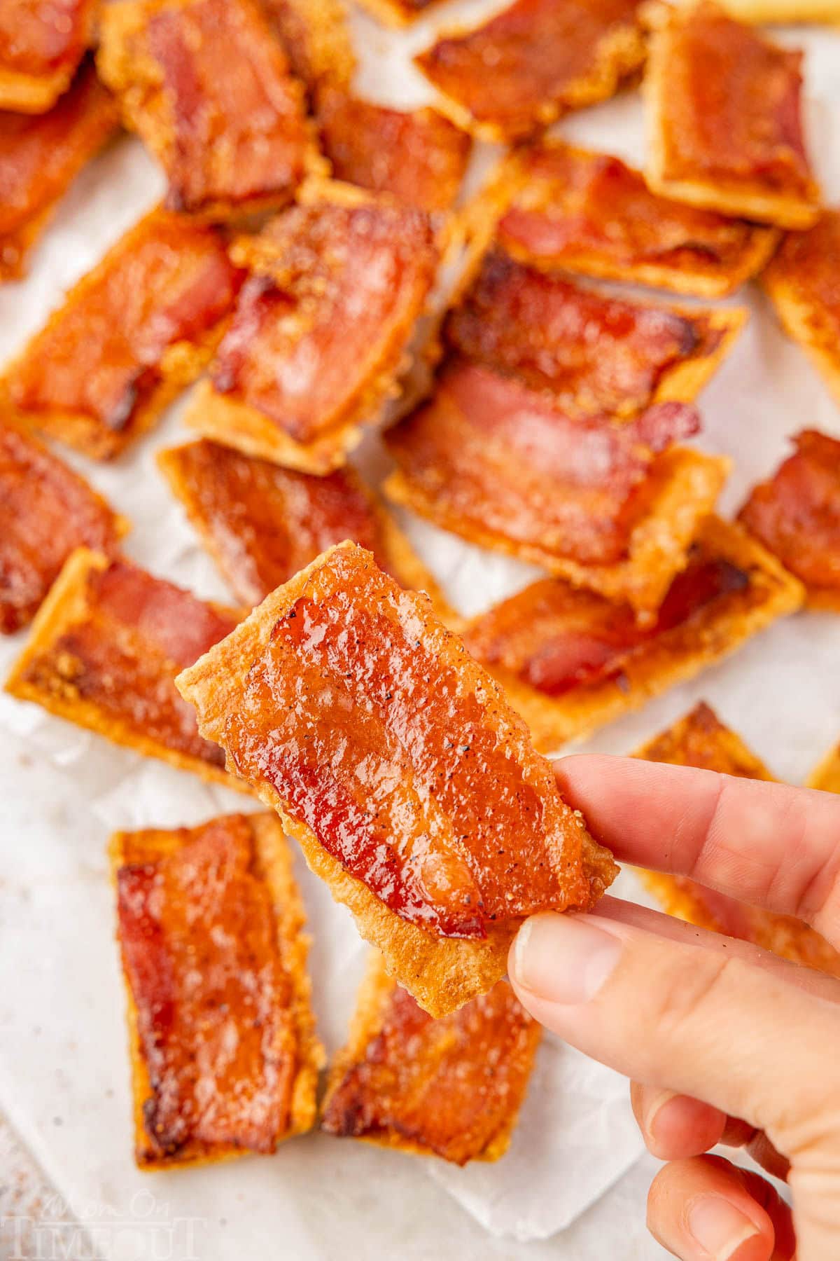 Bacon cracker being held above more crackers on white parchment paper.