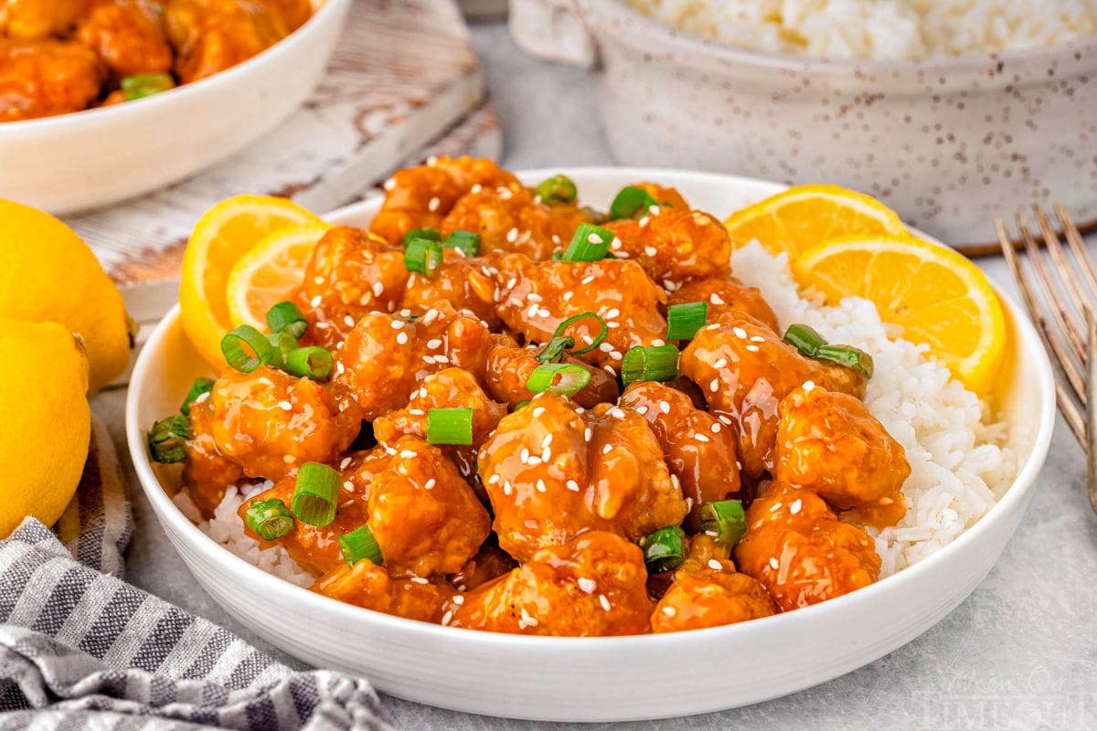 Wide shot of chinese lemon chicken reicpe on white round plate served with white rice and garnished with lemon slices.