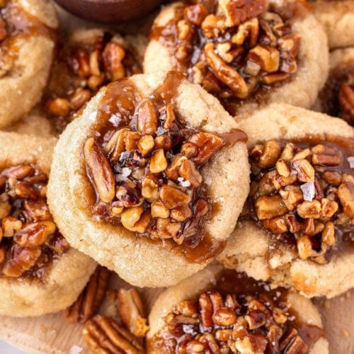Overhead shot of pecan pie cookies piled high on a wood board next to a small bowl of flaky sea salt.