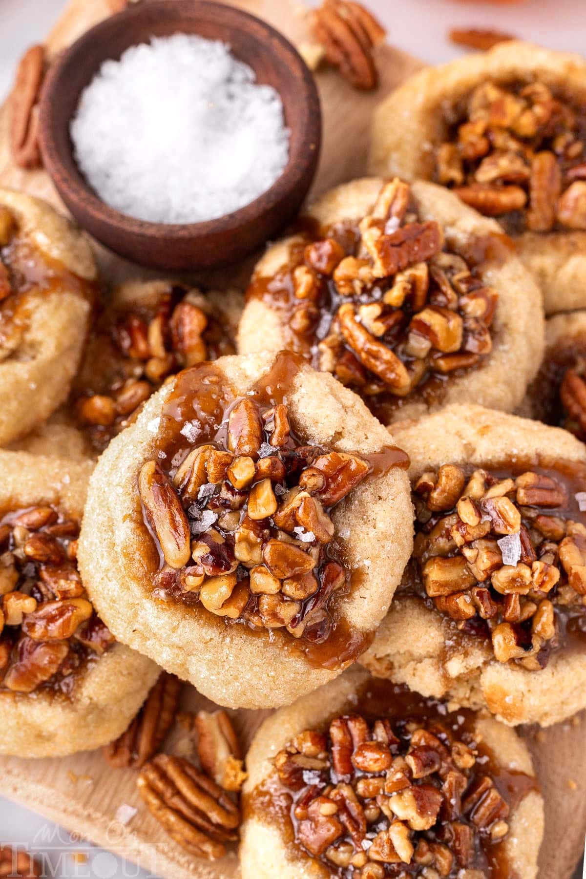 Overhead shot of pecan pie cookies piled high on a wood board next to a small bowl of flaky sea salt.