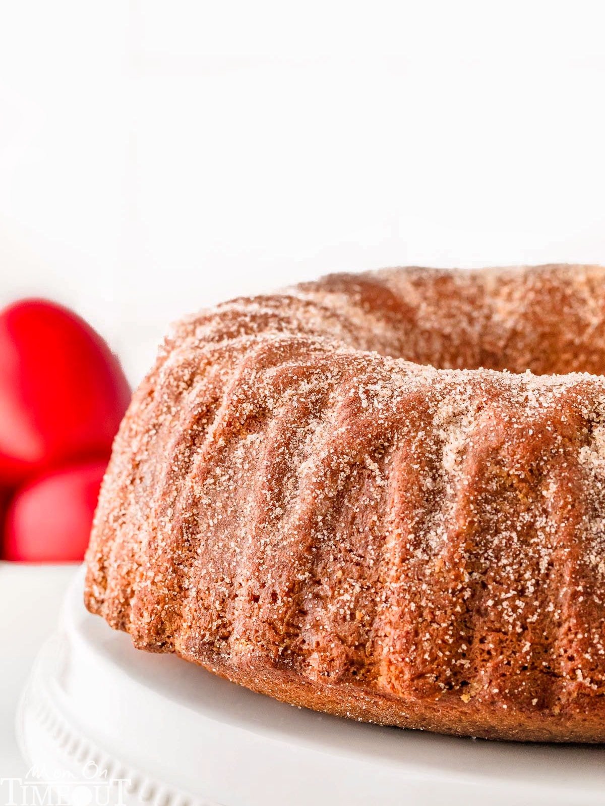 Side view of whole apple cider bundt cake on white cake stand with red apples in the background.