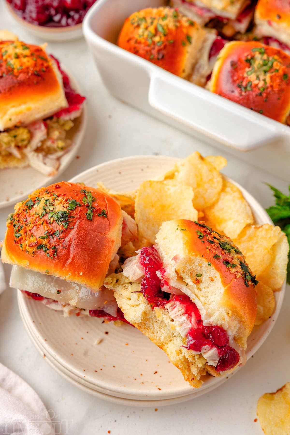 Two turkey sliders on white plate with chips. White baking dish is in the background with the remaining sliders. One slider has a bite taken out of it.