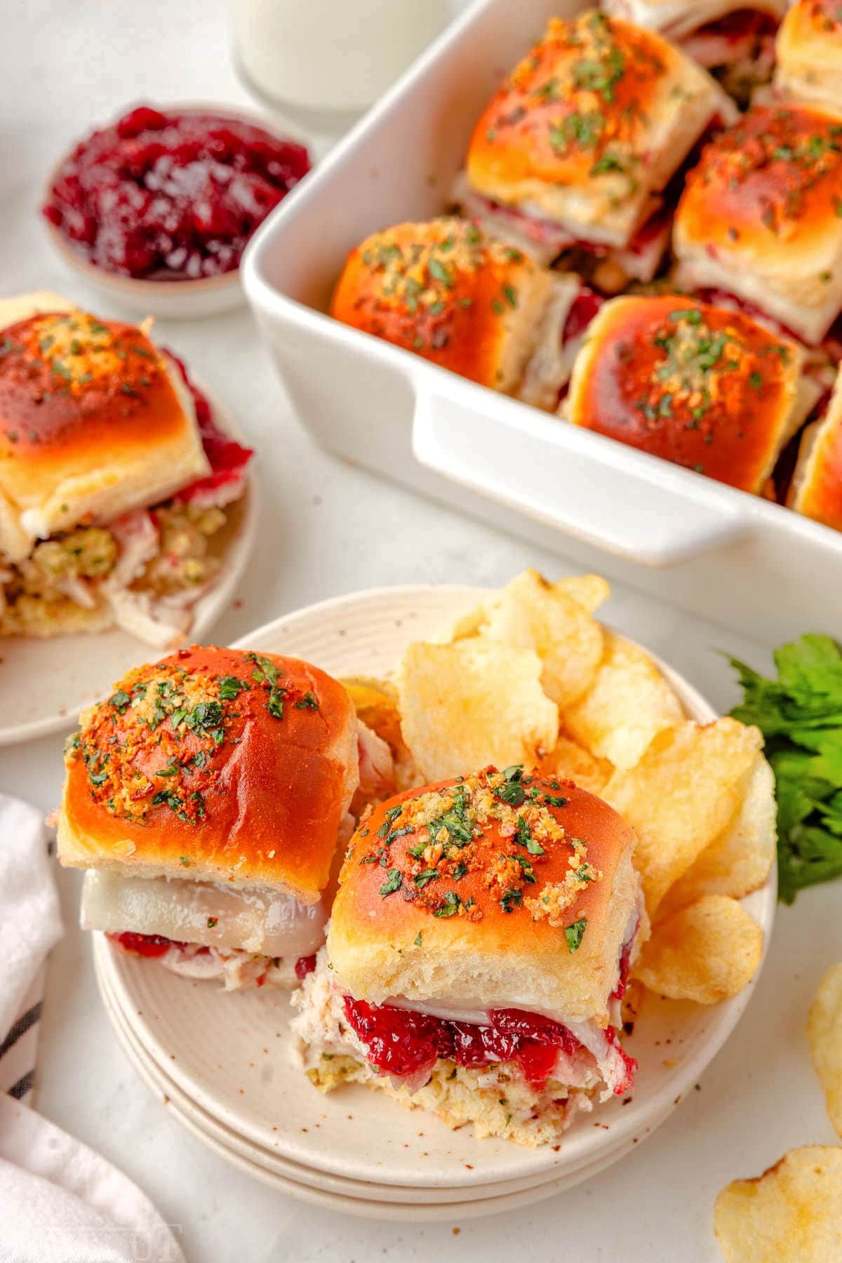 Two turkey sliders on white plate with chips. White baking dish is in the background with the remaining sliders.