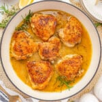 Overhead shot of large white skillet with braised chicken thighs and rosemary and thyme sprigs in the pan.