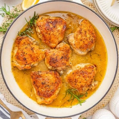 Overhead shot of large white skillet with braised chicken thighs and rosemary and thyme sprigs in the pan.
