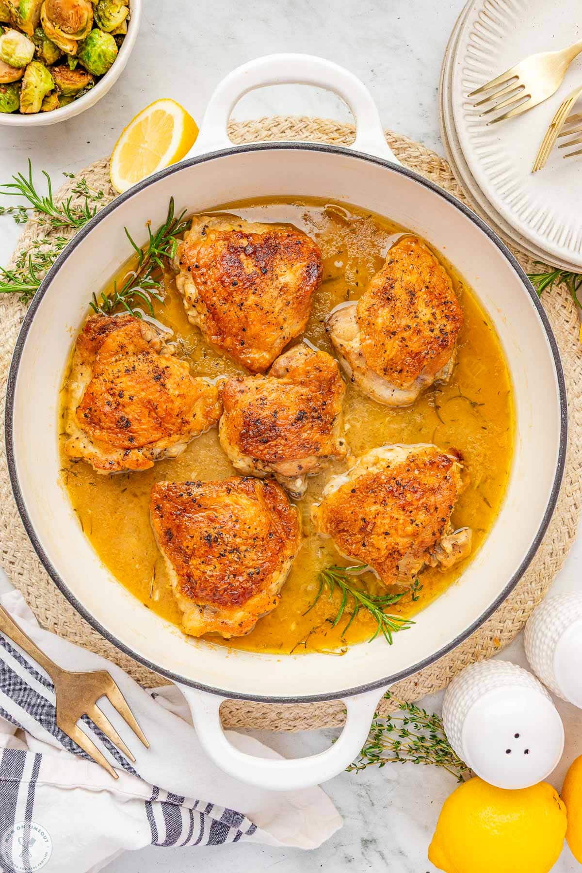 Overhead shot of large white skillet with braised chicken thighs and rosemary and thyme sprigs in the pan.