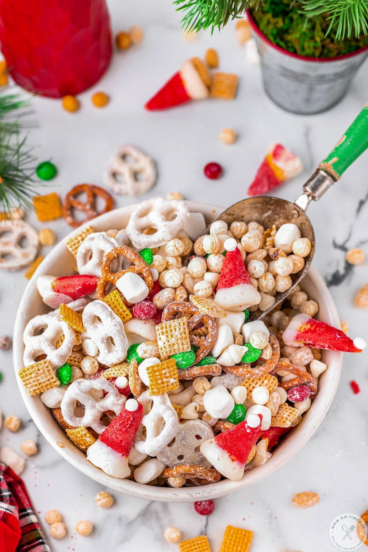 White bowl filled with christmas snack mix with small scoop. Pretzels, peanuts and candy are scattered about the bowl.