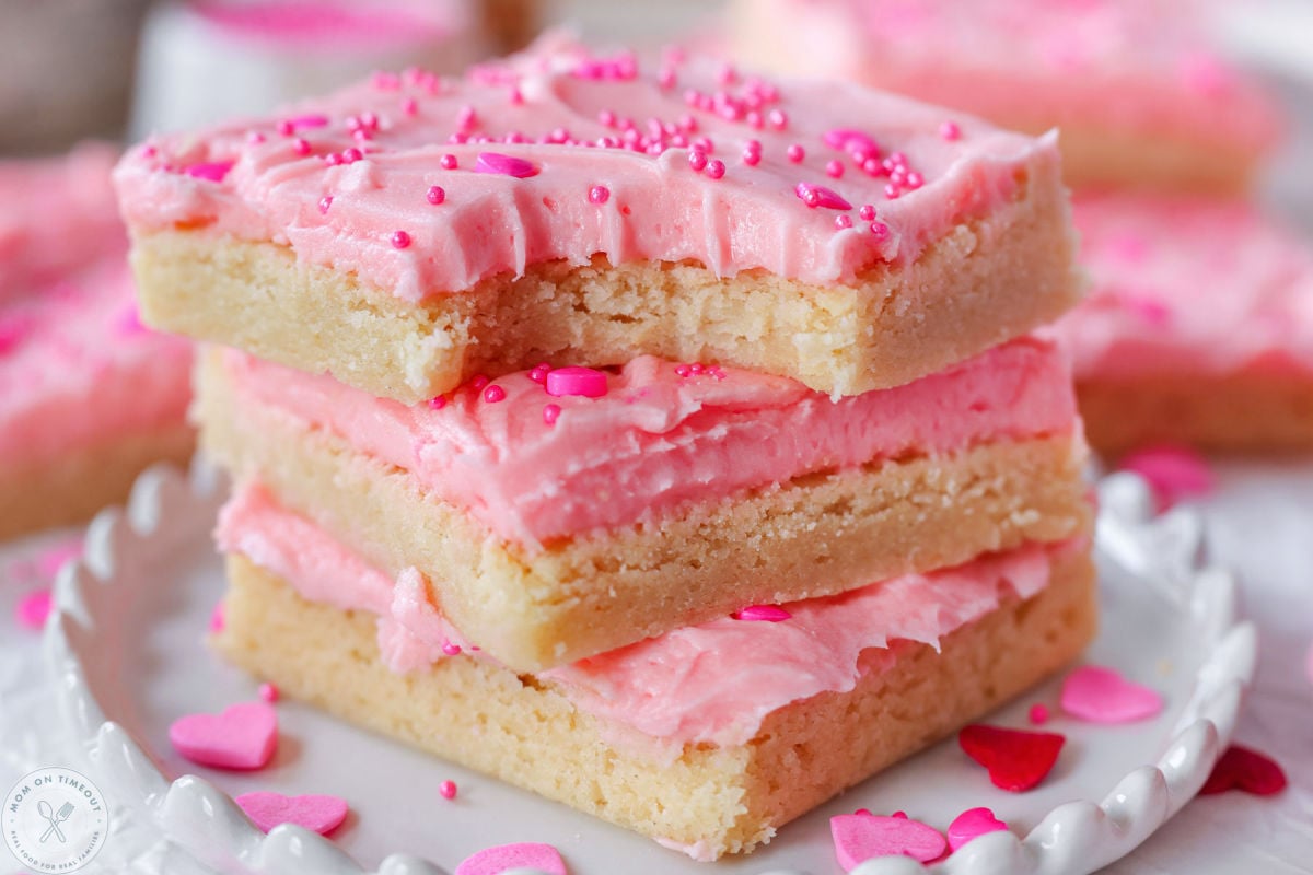 Close up shot of valentines day cookie bars stacked three high on round white scalloped edge plate. Top bar has a bite taken.