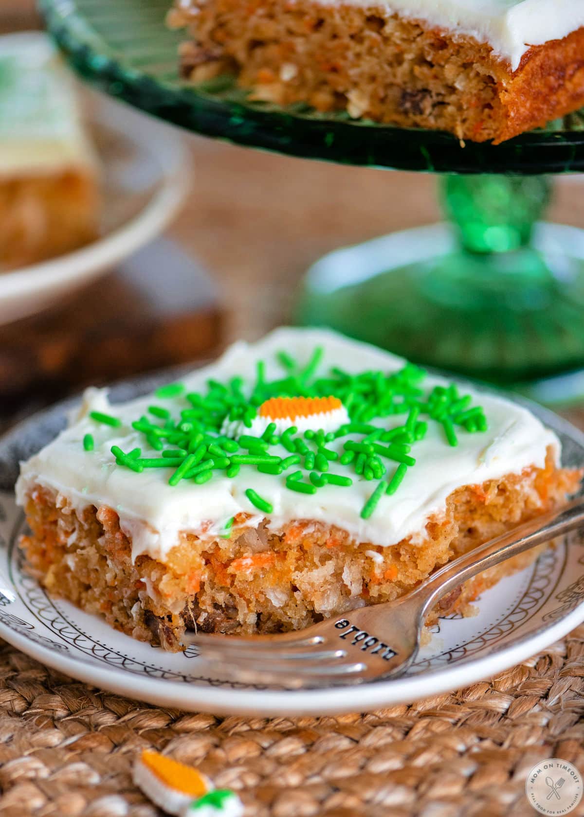 Carrot cake bar on white round plate with bite taken. Fork is sitting on the plate. Cream cheese frosting on top with green sprinkles.