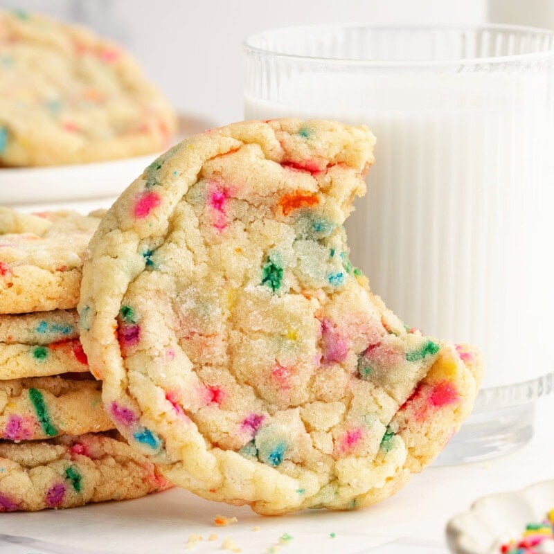 Single funfetti cookie with bite out of it sitting in front of glass of milk.