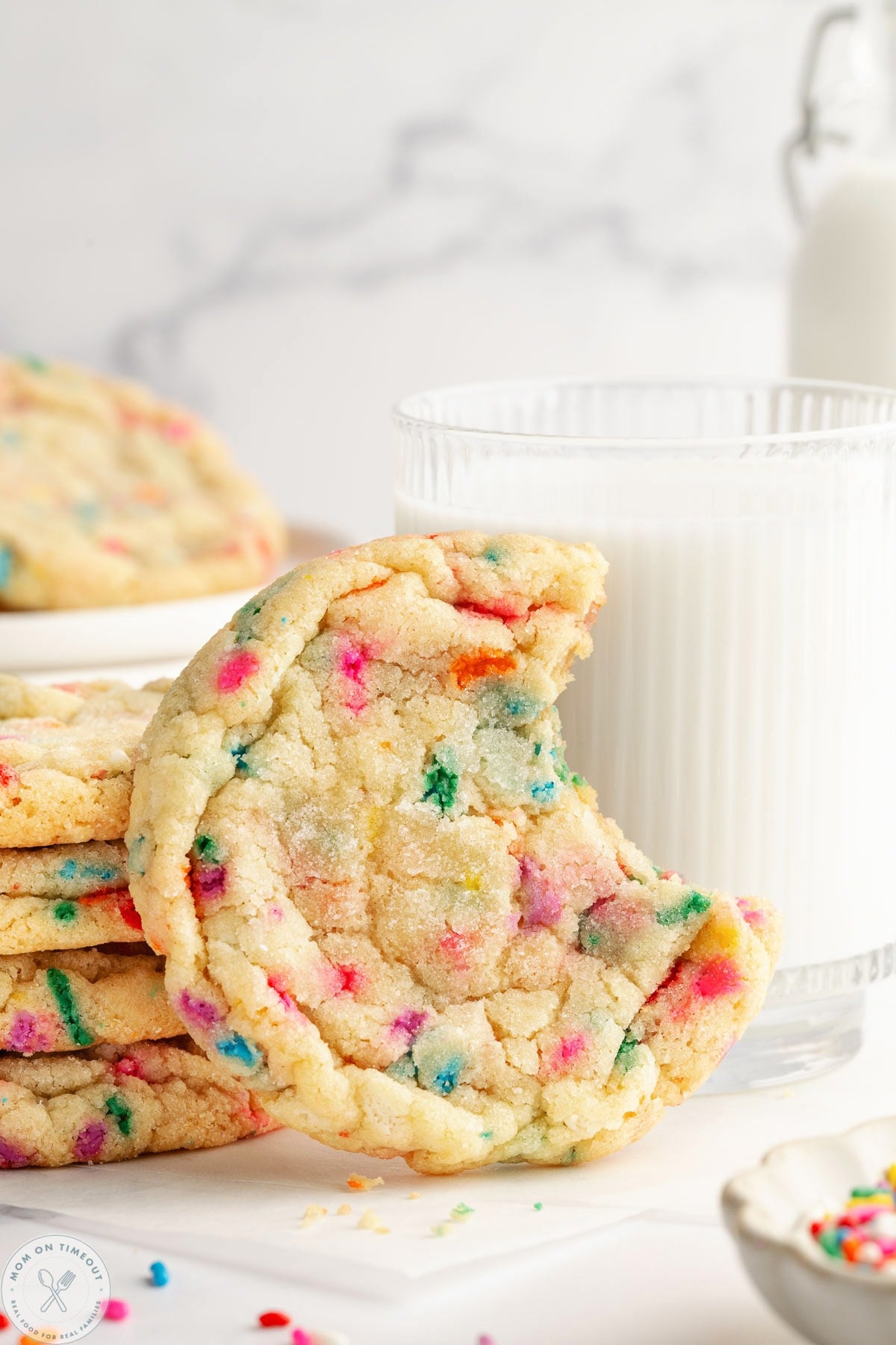 Single funfetti cookie with bite out of it sitting in front of glass of milk.