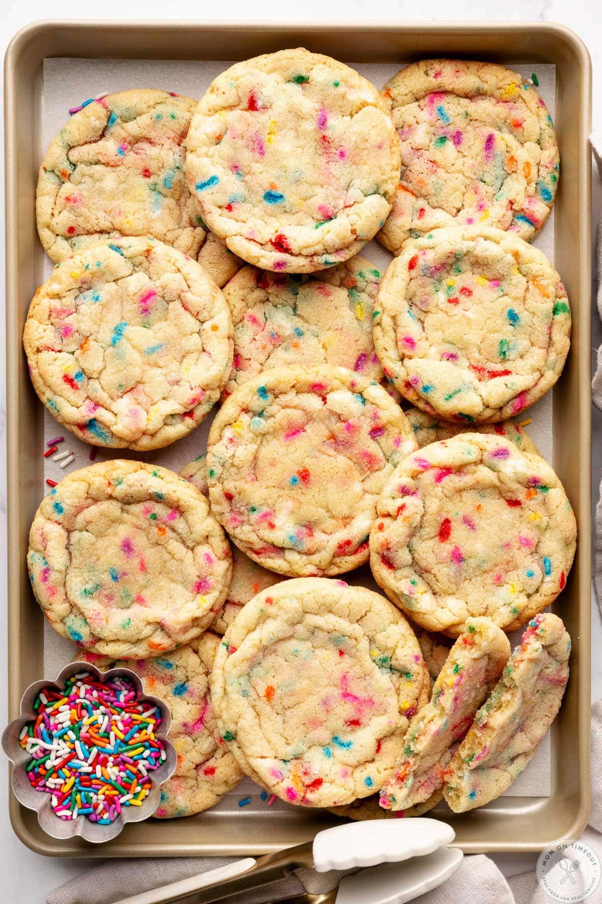 Sprinkle cookies on gold baking sheet with small bowl of jimmies sitting next to the cookies.