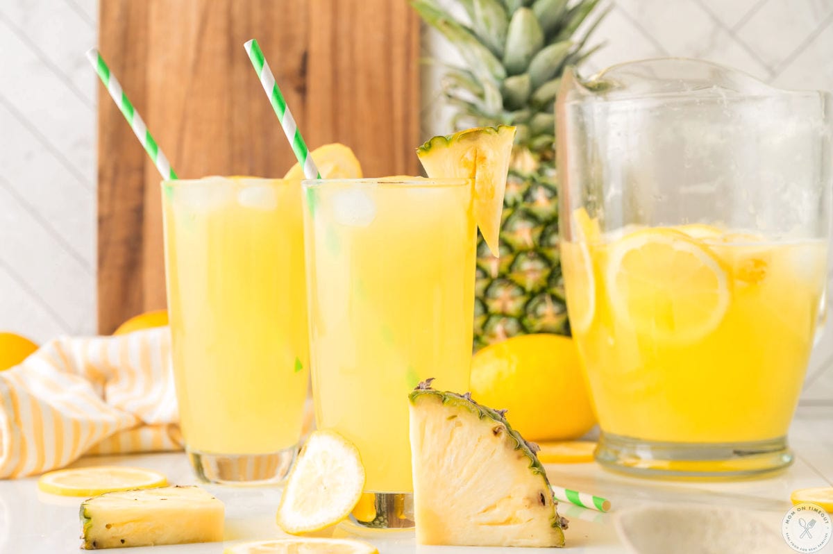 Two glasses filled with pineapple lemonade. Straws in glasses with fresh pineapple in the background.