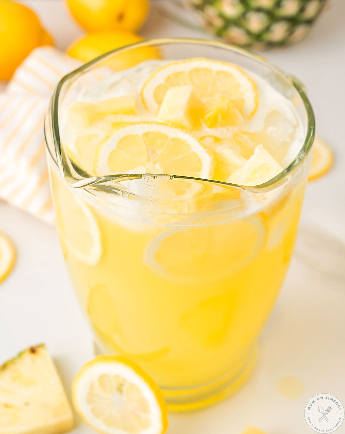 Angled overhead shot of pineapple lemonade recipe in glass pitcher garnished with lemon slices and pineapple chunks.