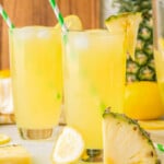 Two glasses filled with pineapple lemonade. Straws in glasses with fresh pineapple in the background.
