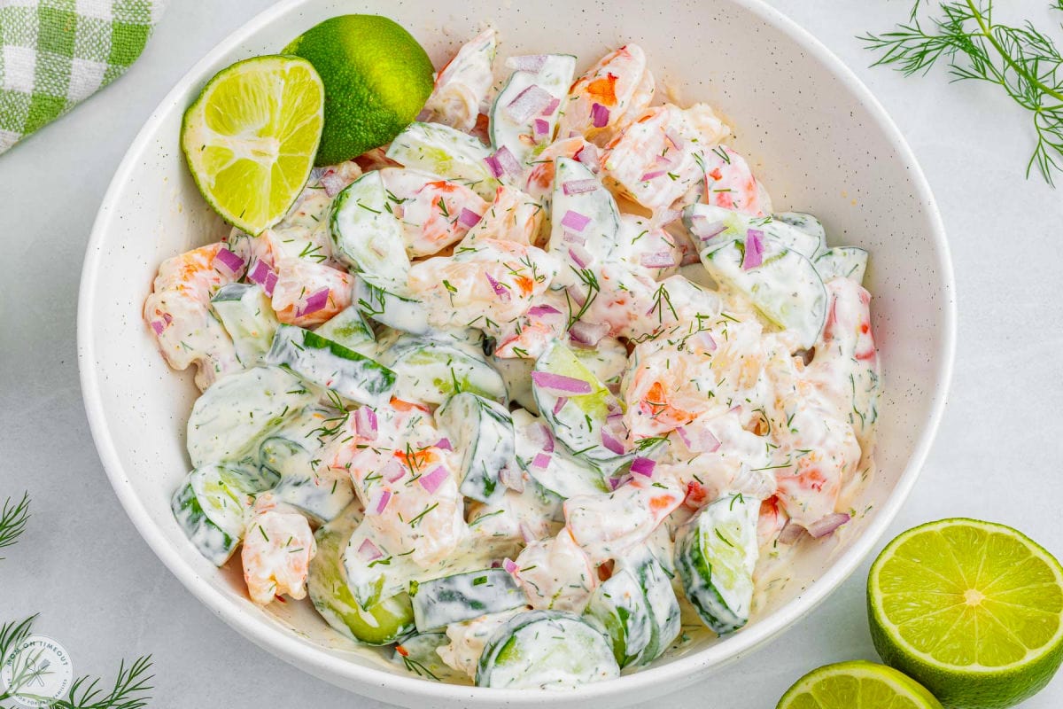 Wide overhead angled shot of cucumber and shrimp salad in a large white serving bowl with two lime wedges off to the side.