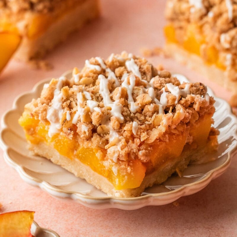 Single peach crumb bar with a vanilla glaze cut into a square sitting on round dessert plate with more bars in the background.