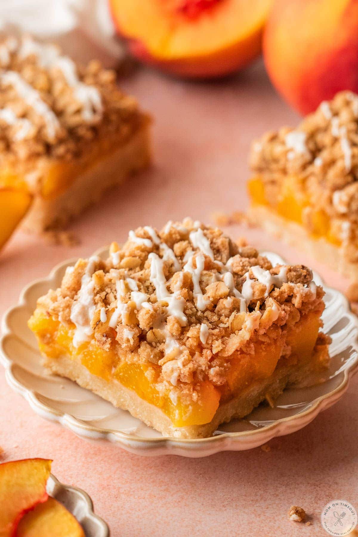 Single peach crumb bar with a vanilla glaze cut into a square sitting on round dessert plate with more bars in the background.