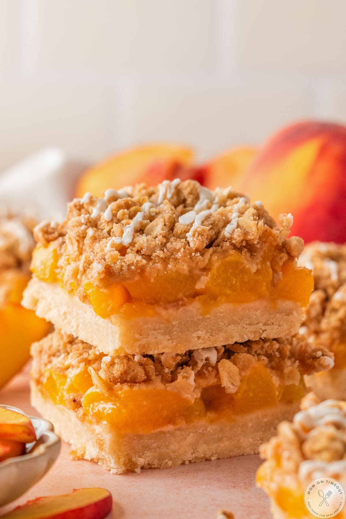 Two whole peach pie crumb bars stacked on each with peaches in the background.