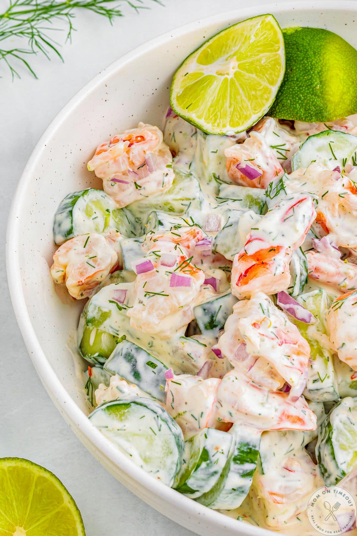 Overhead shot of half of white bowl filled with shrimp and cucumber salad. Two lime wedges are tucked into the salad.