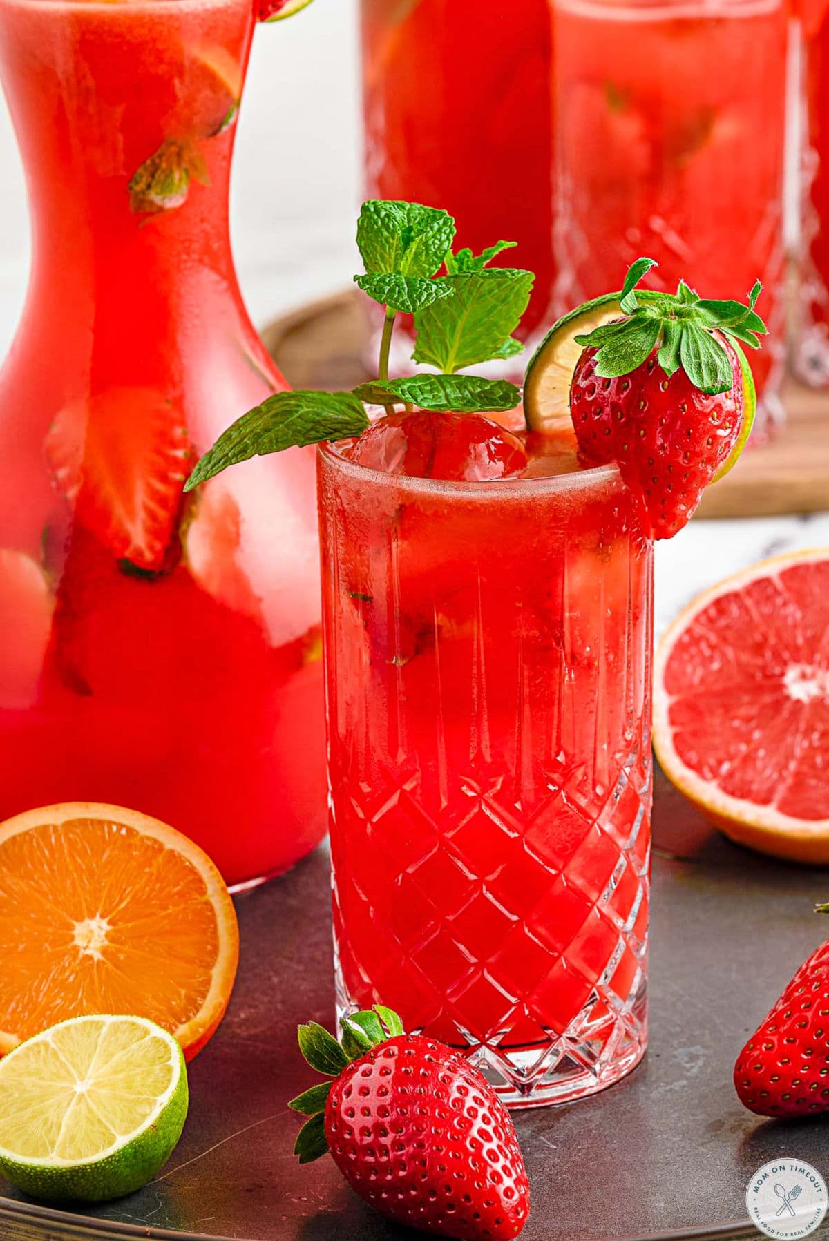 Strawberry mojito drink in a glass garnished with strawberries and fresh mint. Pitcher of mojitos in the background.