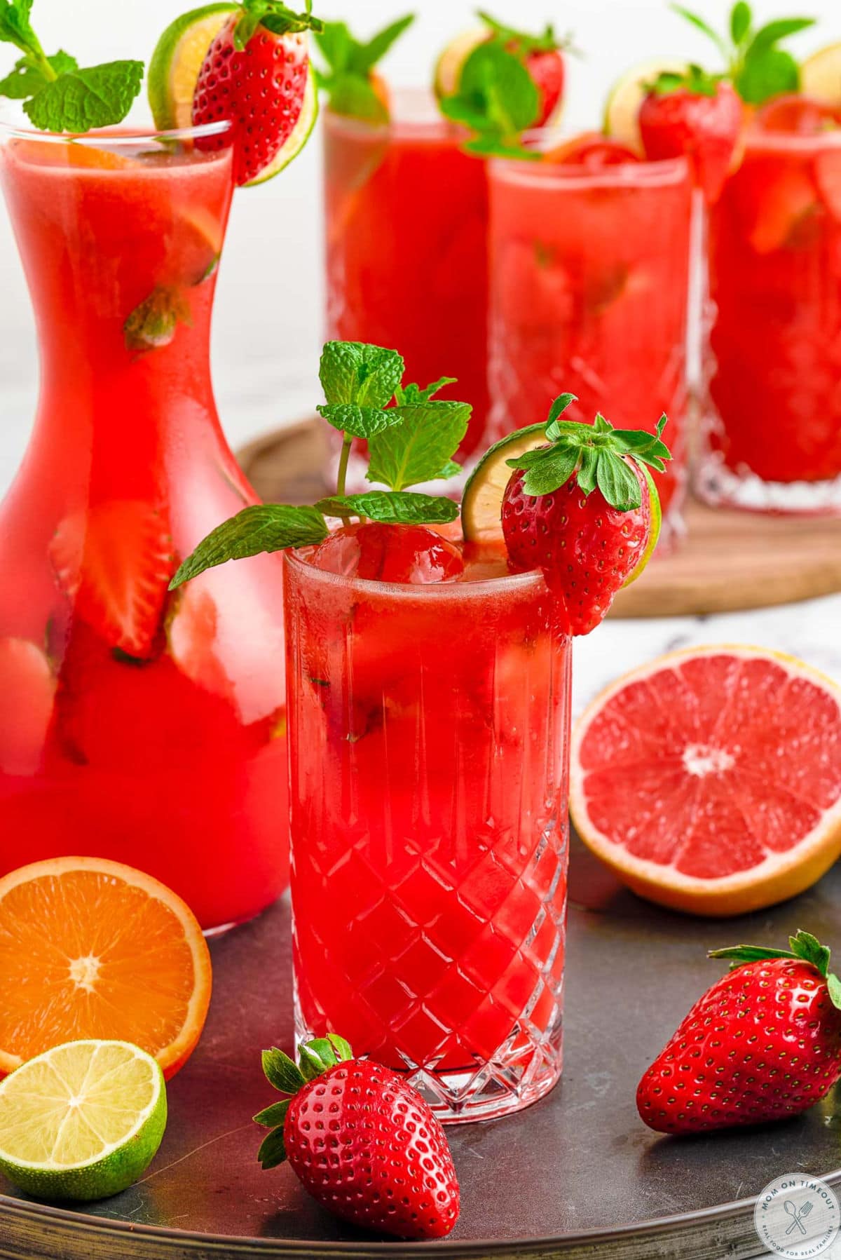 Pulled back shot of glass of strawberry mojito drink with three more glasses filled and a pitcher in the background. Orange slices, lime slices and strawberries are scattered about.