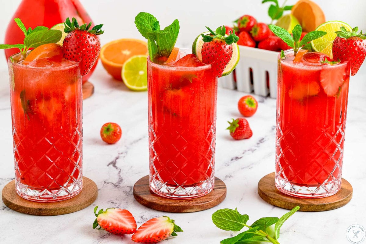 Wide shot of three strawberry mojitos in tall glasses garnished with lime slices, mint, and strawberries.