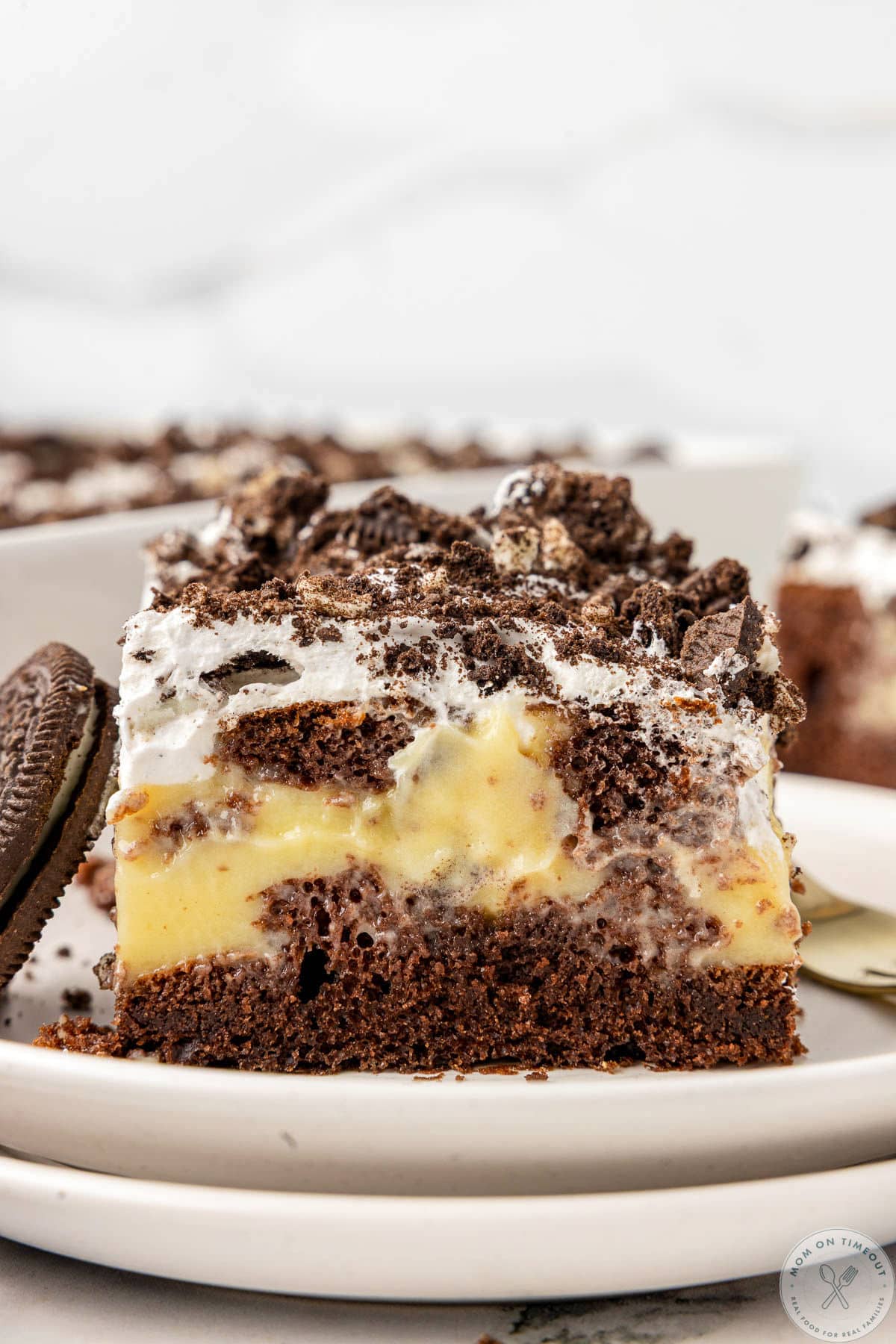 Piece of oreo poke cake on white round plate. Baking dish with the rest of the cake can be seen in the background.