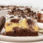 Piece of oreo poke cake on white round plate. Baking dish with the rest of the cake can be seen in the background.