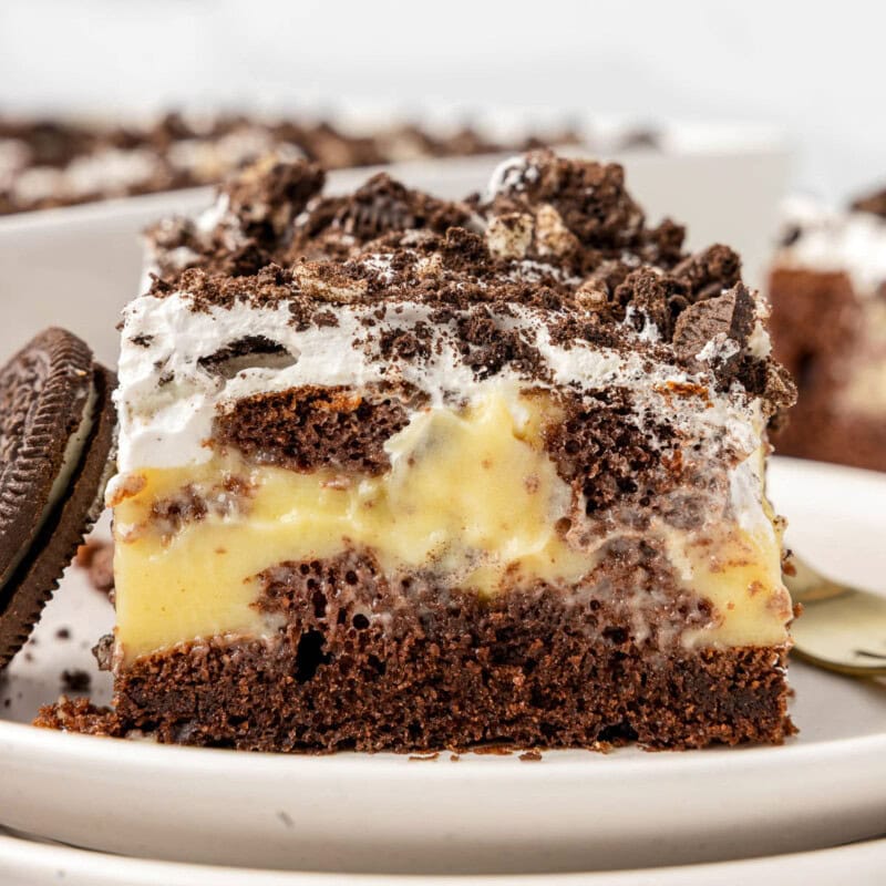 Piece of oreo poke cake on white round plate. Baking dish with the rest of the cake can be seen in the background.