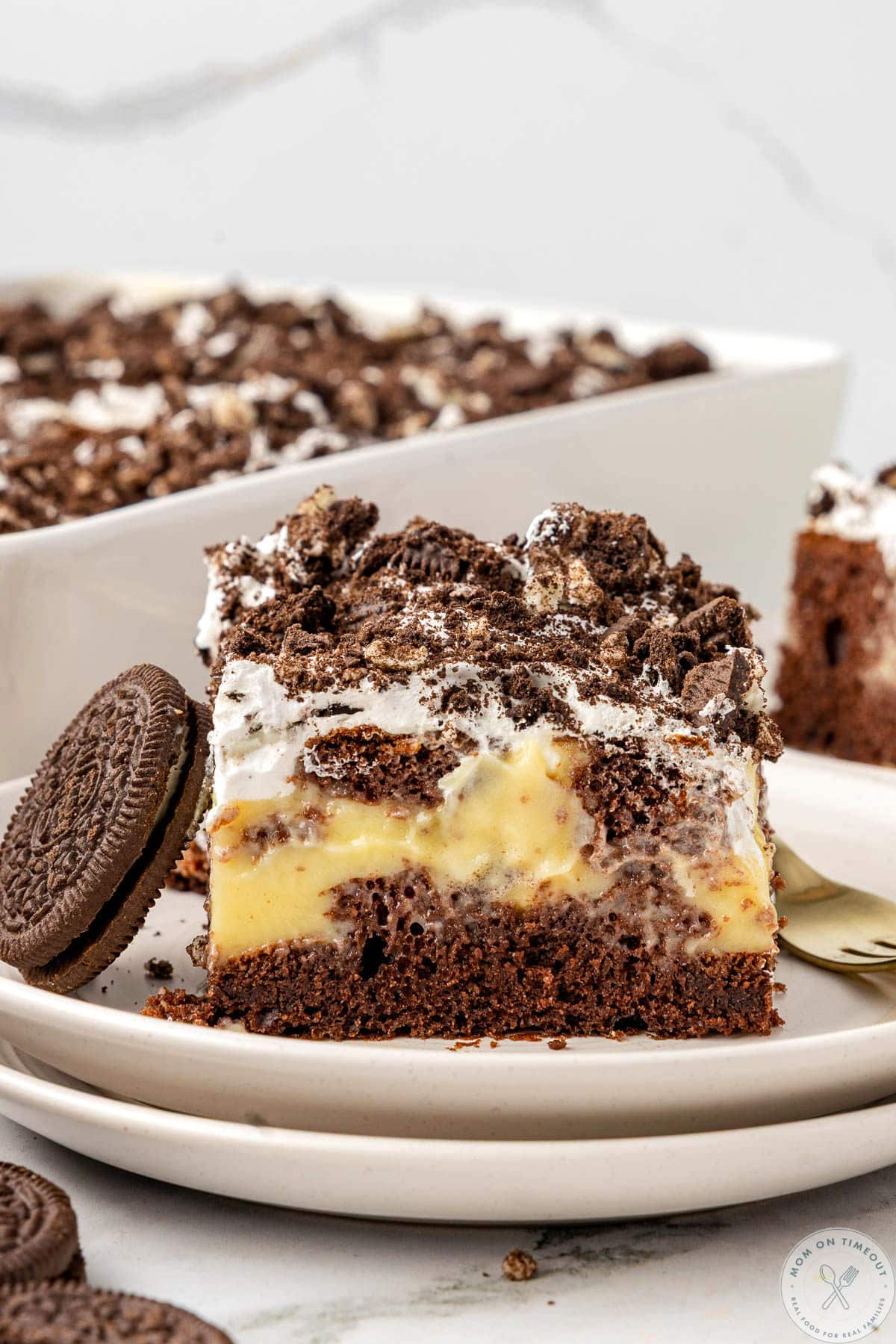 Piece of oreo poke cake on white round plate. Baking dish with the rest of the cake can be seen in the background.